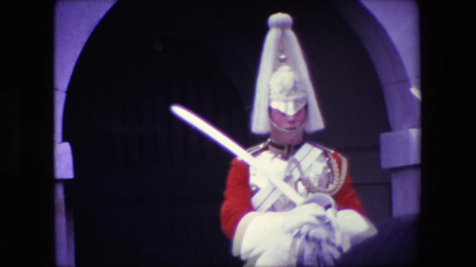 1967 London home-movie frame of a ceremonial mounted rider in red uniform and plumed helmet holding a sword beneath a stone archway.