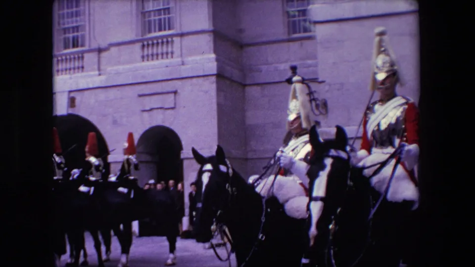 1967 London frame showing a formation of mounted Royal Guards in ceremonial helmets and red uniforms riding past arched stone entrances.