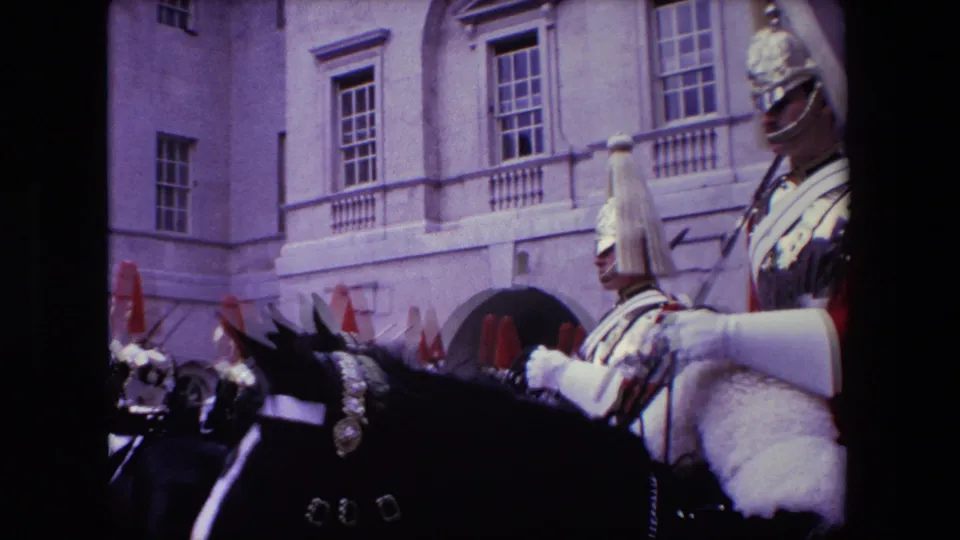 1967 London home-movie frame with mounted Royal Guards and horses passing close to the camera before a pale stone facade with tall windows.