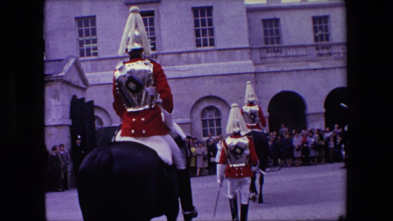 1967 London home-movie frame showing mounted ceremonial soldiers in red uniforms riding past spectators outside a large stone building with arched openings.