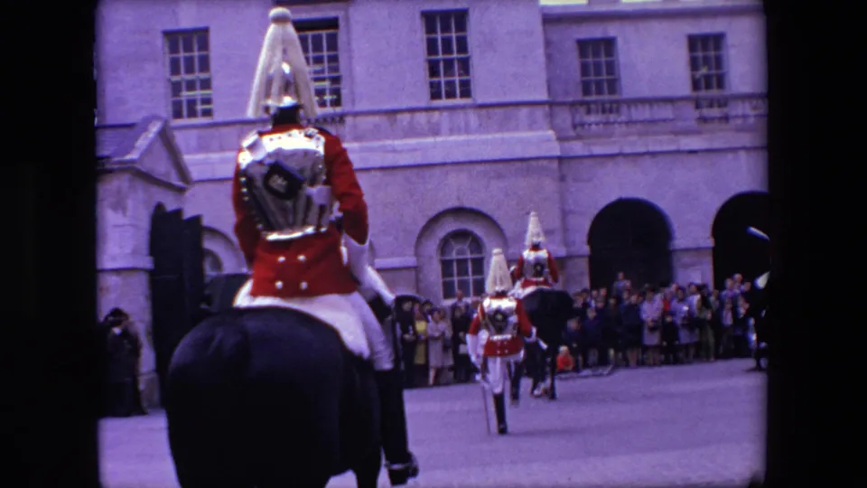 1967 London home-movie view from behind a mounted ceremonial soldier, with additional guards, dark horses, and spectators gathered outside a stone building.