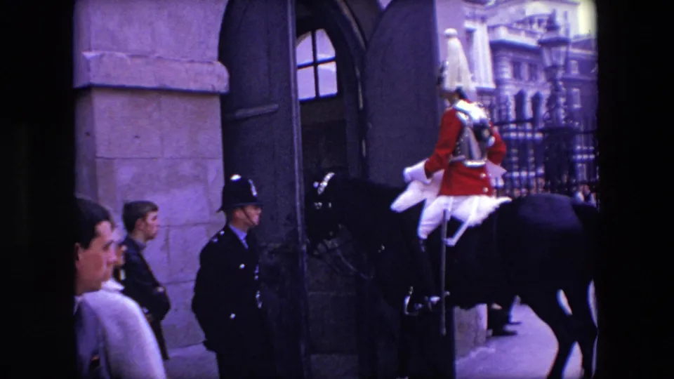 1967 London frame showing a mounted ceremonial guard riding near an exterior wall, a uniformed police officer, pedestrians, and wrought-iron gates.