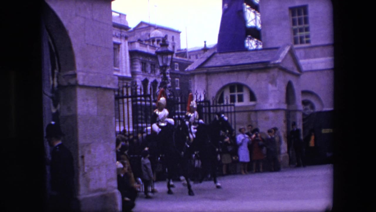 Two mounted armored riders pass in front of iron railings and a crowd outside stone buildings.
