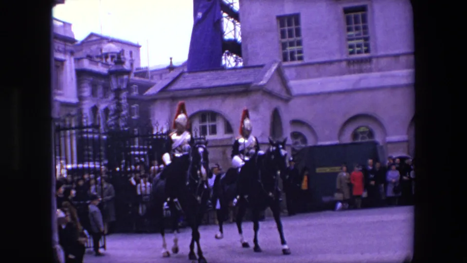 1967 London home-movie frame showing two armored ceremonial riders on horseback crossing a paved area before a crowd gathered near iron railings.
