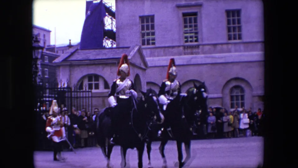 1967 London frame showing mounted ceremonial riders in armor moving in formation past onlookers beside stone buildings and iron fencing.