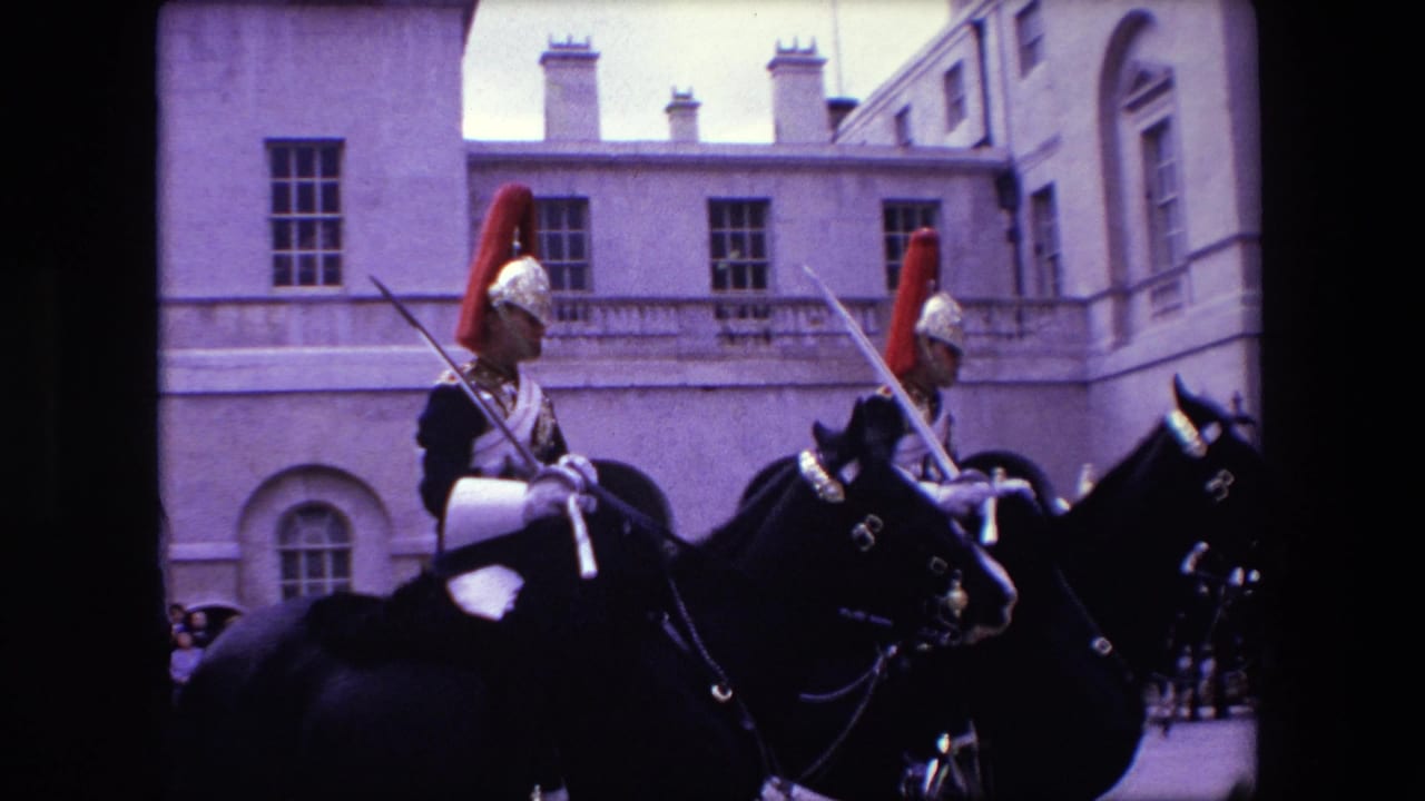 Two mounted soldiers with swords ride close to the camera in front of stone buildings.