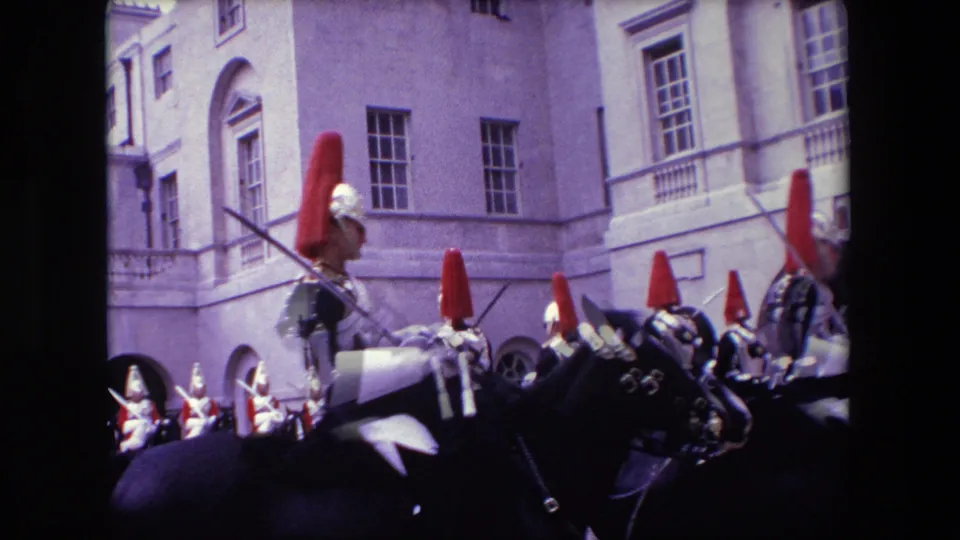 A group of mounted ceremonial soldiers with red plumes and polished armor ride or gather in formation near pale stone architecture, with additional uniformed figures partly visible at the edge of the frame.