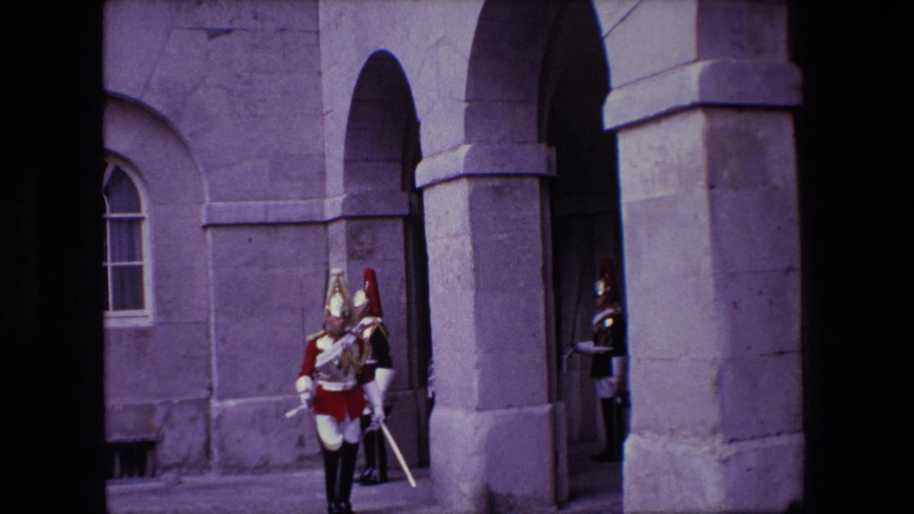 1967 London home-movie frame showing ceremonial soldiers in ornate helmets and plumed headgear near a stone arched entrance.