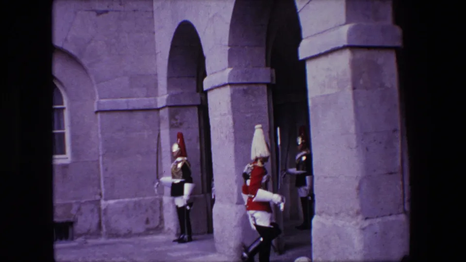 1967 London home-movie frame showing a white-uniformed ceremonial soldier moving past guards at a stone arched doorway.