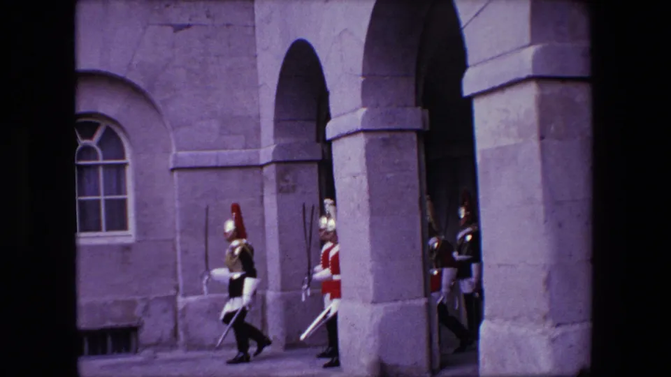 1967 London home-movie frame with uniformed guards moving along a stone building facade with arched openings.