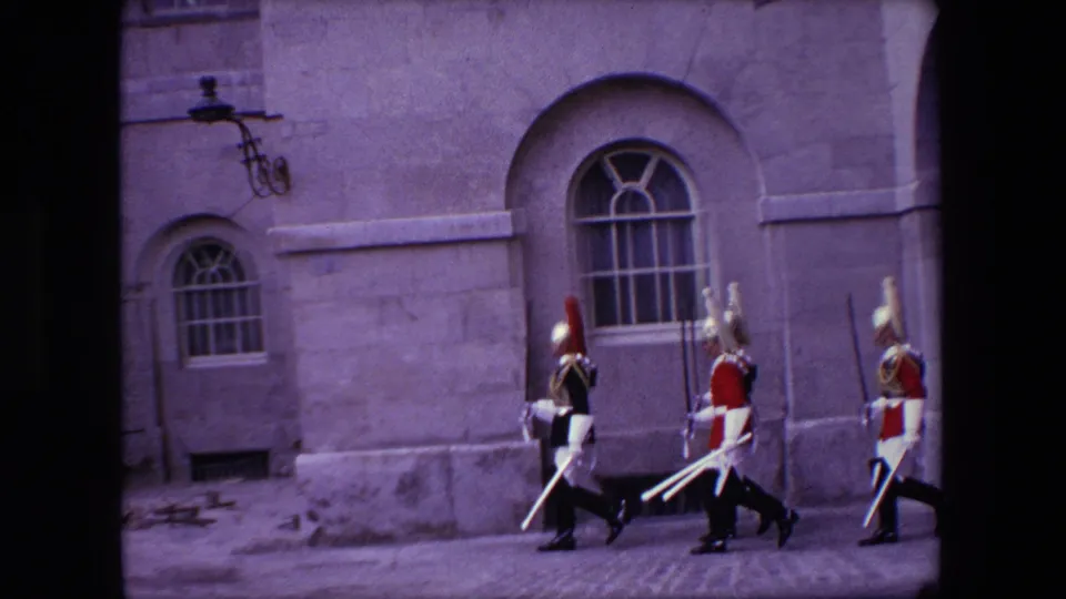1967 London frame showing three ceremonial uniformed guards walking past a stone facade with arched windows.