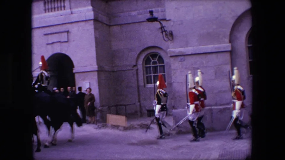 1967 London 8mm frame with uniformed guards, a mounted figure partly visible, and spectators near a stone building entrance.
