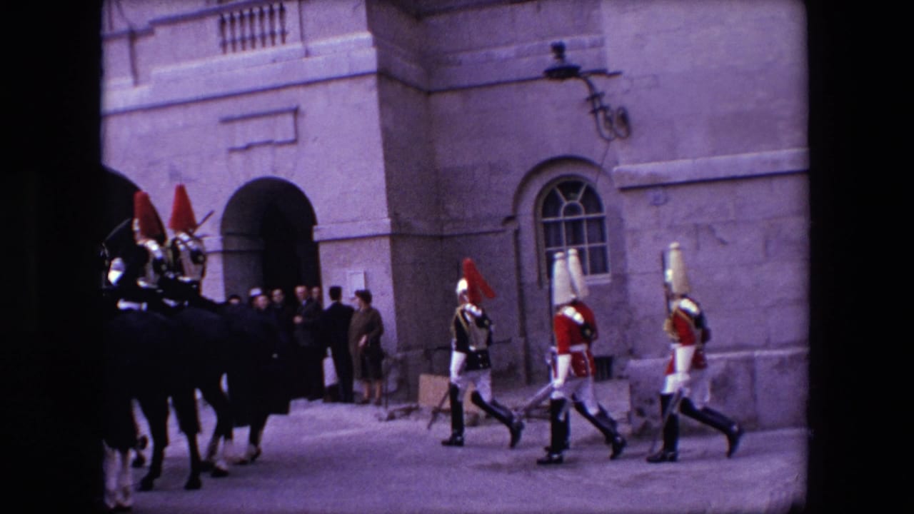 1967 London 8mm frame showing mounted guards and marching ceremonial soldiers moving past a stone facade as a small group of spectators watches from near an arched entrance.
