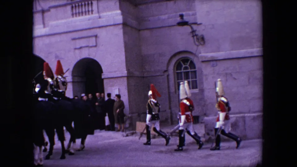 1967 London 8mm frame showing mounted guards and marching ceremonial soldiers moving past a stone facade as a small group of spectators watches from near an arched entrance.
