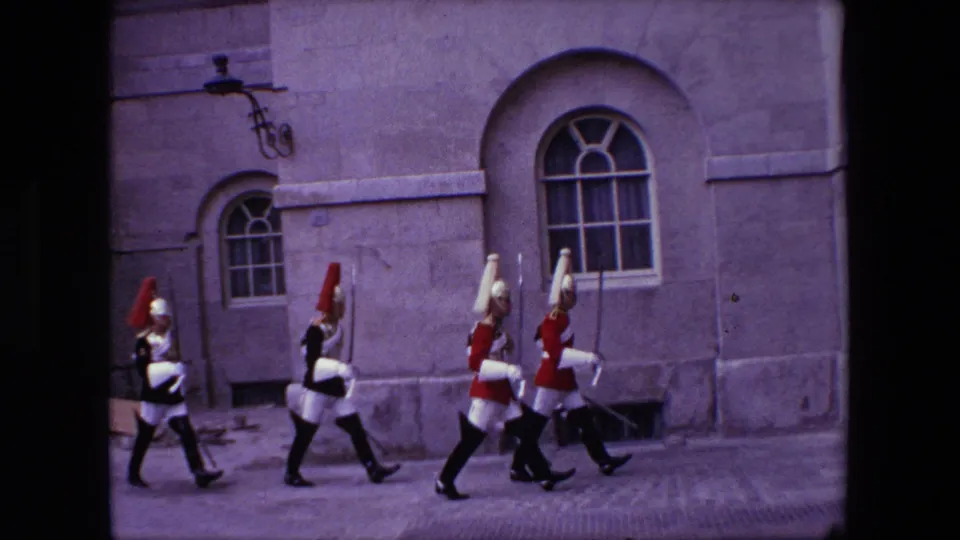 1967 London 8mm frame showing ceremonial soldiers in red and dark uniforms marching along a paved area beside arched windows in a large stone building.