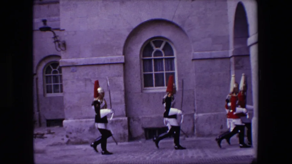 1967 London 8mm frame showing uniformed ceremonial guards continuing past a stone facade with arched windows, framed by dark film edges from the scan.
