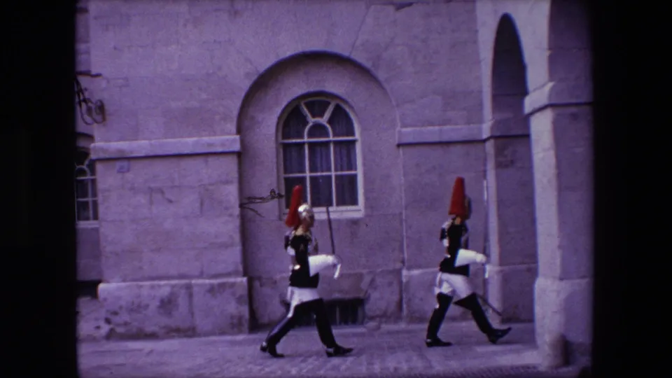Two ceremonial soldiers in dark uniforms and red-plumed helmets march across a London stone courtyard in 1967 home-movie footage.