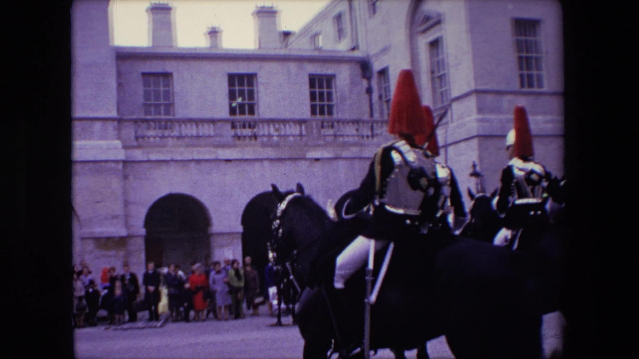 In London 1967 home-movie footage, mounted ceremonial guards with red plumes ride past a pale stone building while a crowd watches from the courtyard edge.
