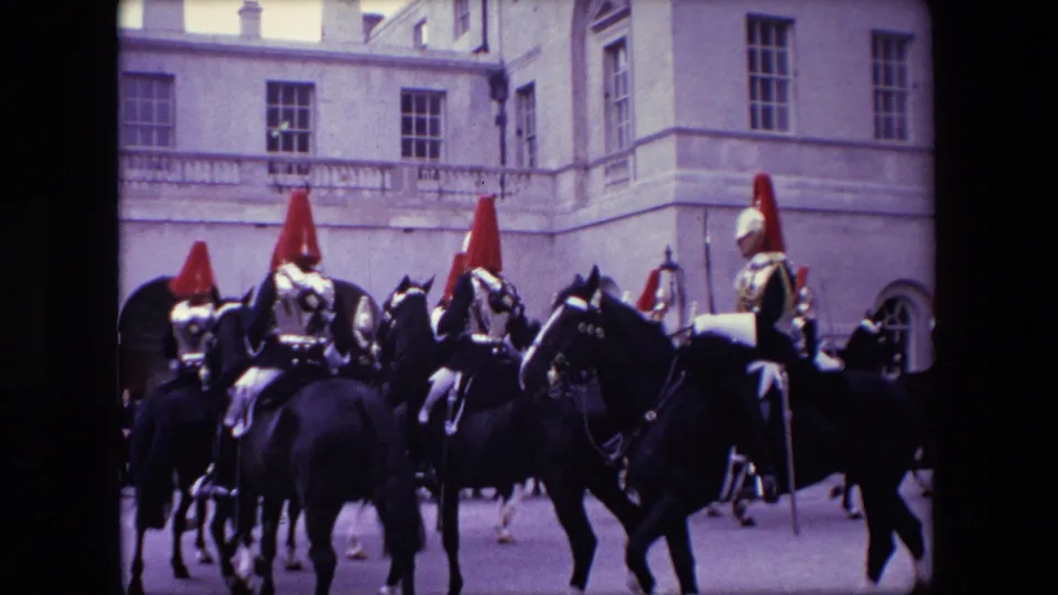 1967 London home-movie frame showing mounted guards with red helmet plumes riding dark horses past formal stone architecture.