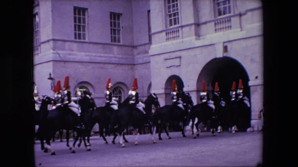 Mounted guards and dark horses move across a London street beside a pale building with tall windows and a large arched opening.