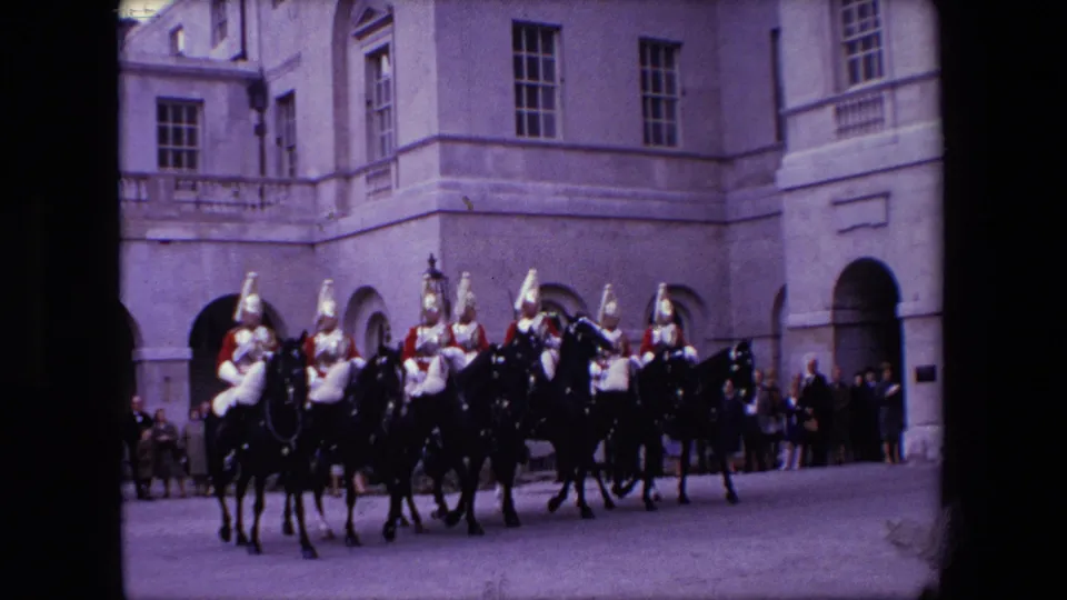 Mounted ceremonial guards in helmets and red or dark uniform elements ride in formation across a paved London forecourt in the 1967 home-movie clip.