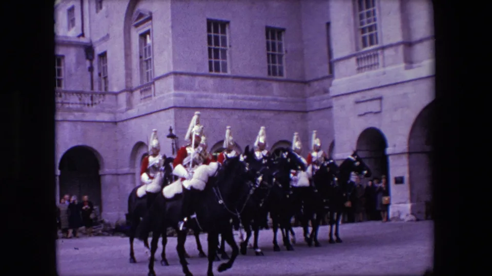 The frame shows mounted ceremonial guards gathered near an arched entrance, with spectators and a pale stone institutional building visible behind them.