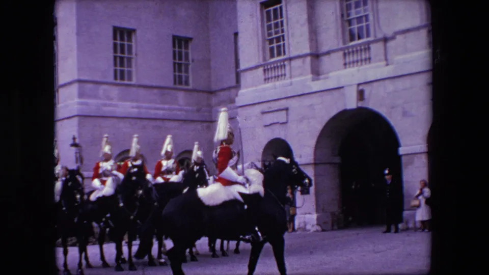A mounted ceremonial guard wearing red uniform elements rides past other mounted personnel near a large arched doorway in the London 1967 archival footage.