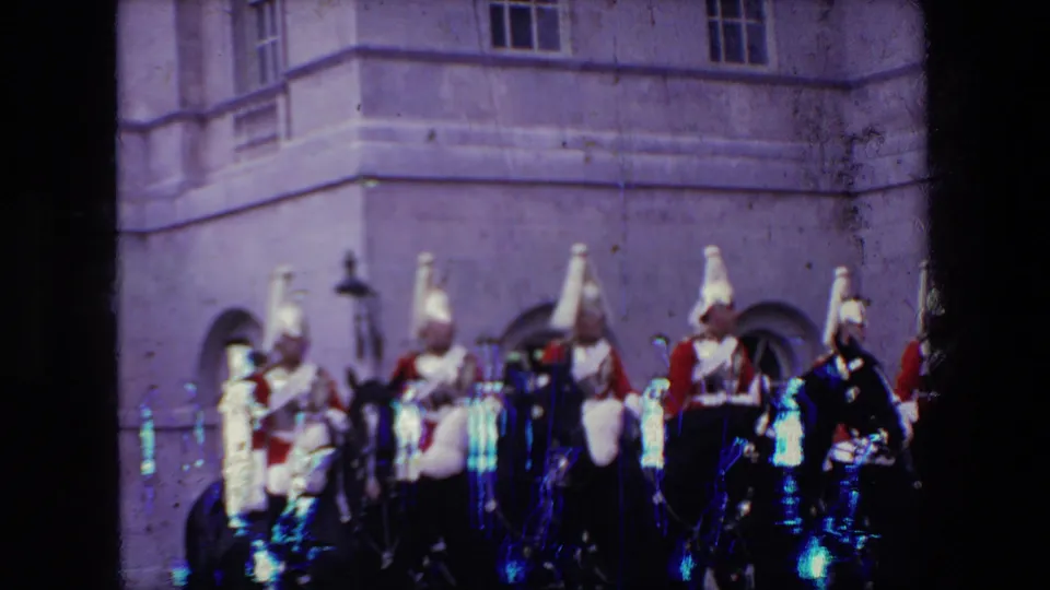 A closer, softer 8mm frame shows mounted uniformed riders and black horses passing the stone building, with visible color shift and film wear.