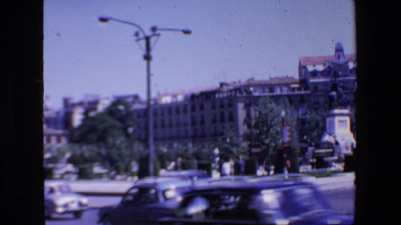 Street traffic passes in front of buildings, trees, a streetlamp, and a monument.