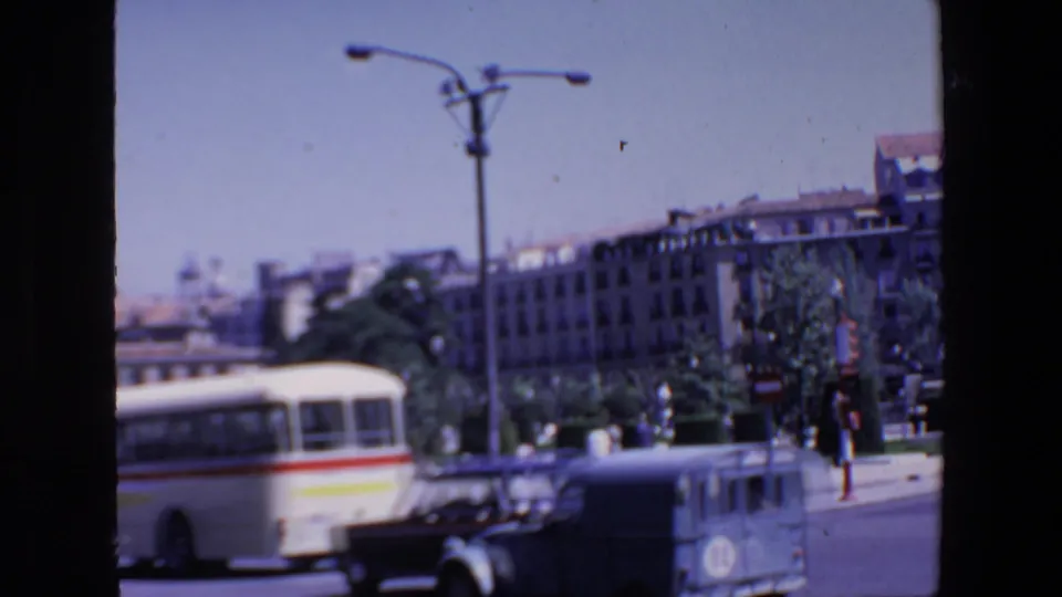 Madrid street traffic in the 1967 home-movie footage, showing a white bus with a red stripe, cars, pedestrians, trees, a streetlamp, and civic buildings near a monument.