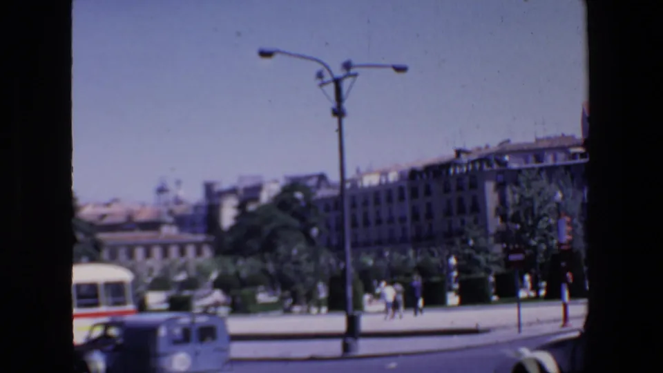 Soft 8mm view of a Madrid public street in 1967, with a bus at the lower edge, pedestrians near the square, trees, a streetlamp, background buildings, and a monument.