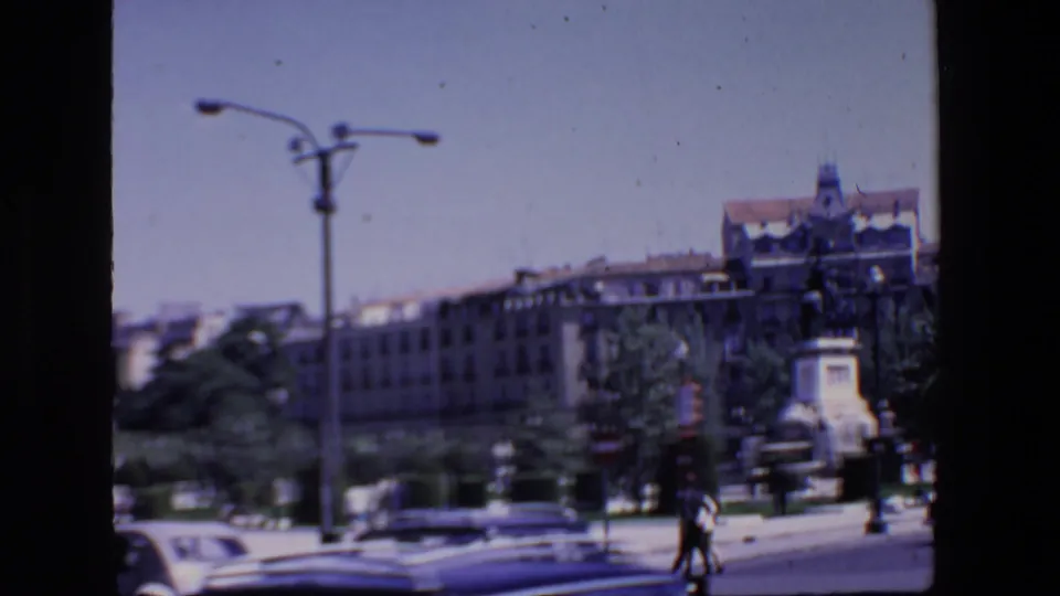 Madrid street view from the 1967 home movie, with pedestrians at the roadside, blurred cars in the foreground, trees, a tall streetlamp, multi-story buildings, and a monument at right.