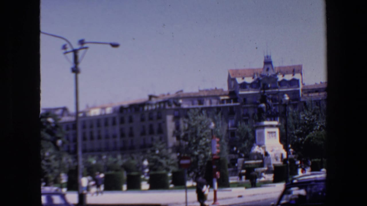 1967 Madrid home-movie frame showing a public monument on a pedestal, surrounding plaza greenery, street lamps, distant pedestrians, vehicles, and large buildings in the background.