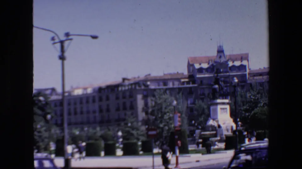 1967 Madrid home-movie frame showing a public monument on a pedestal, surrounding plaza greenery, street lamps, distant pedestrians, vehicles, and large buildings in the background.