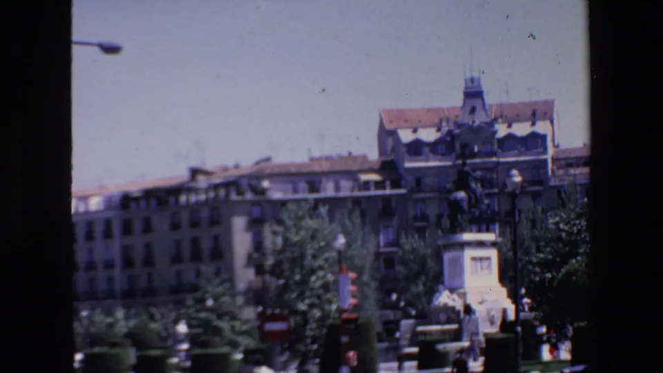1967 Madrid frame with a mounted public monument, stone base, formal plantings, street lamps, distant figures, and multi-story buildings under bright daylight.