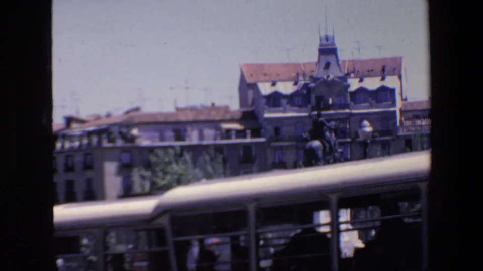 1967 Madrid home-movie frame with a bus crossing the foreground, partially blocking a plaza monument, with street lamps, trees, and older city buildings behind it.