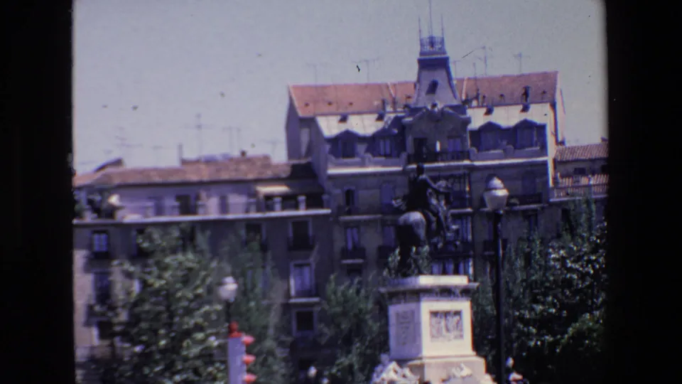 1967 Madrid frame showing a mounted public monument and pedestal more prominently, with leafy trees, street lamps, distant pedestrians, and urban buildings in the background.