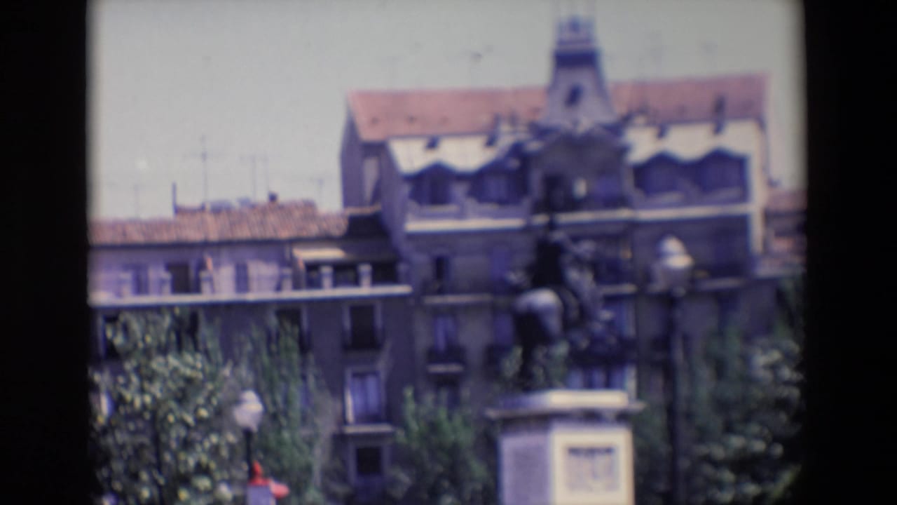 Soft 1967 Madrid home-movie frame showing an equestrian statue on a pedestal with apartment buildings, trees, and film borders around the view.