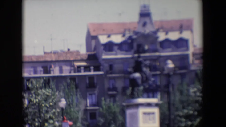 Soft 1967 Madrid home-movie frame showing an equestrian statue on a pedestal with apartment buildings, trees, and film borders around the view.