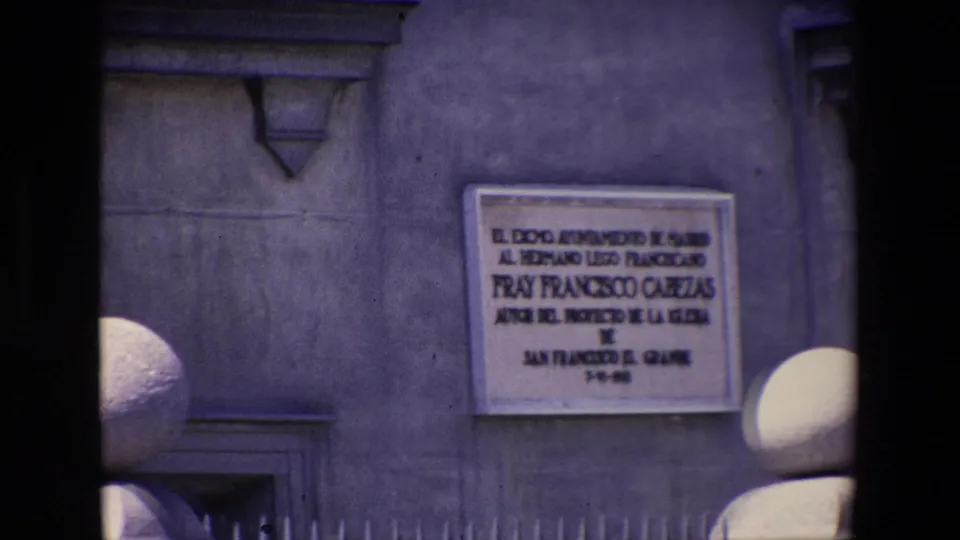 1967 Madrid home-movie frame focused on a Spanish commemorative plaque set into a stone wall, with globe lamps and historic facades in the background.