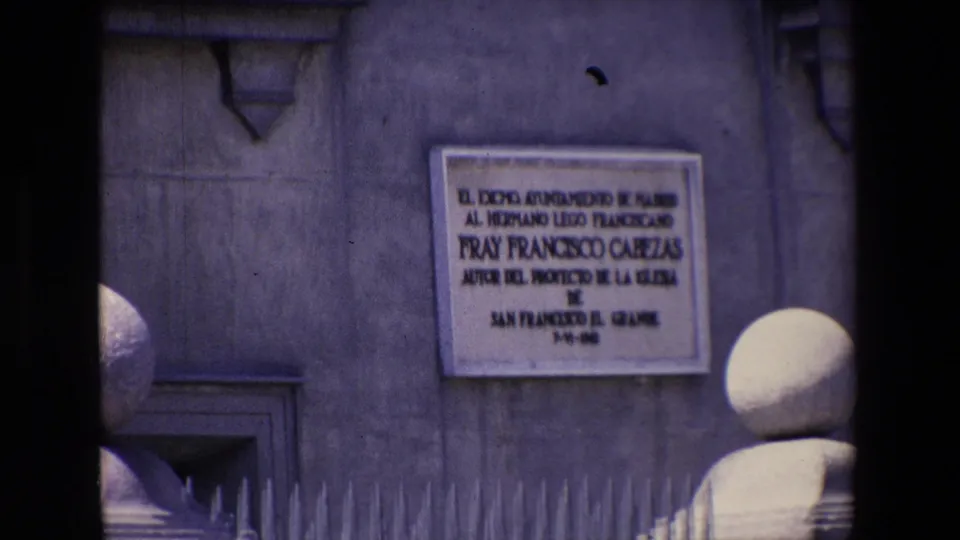 1967 Madrid home-movie frame showing a weathered stone facade with a large Spanish text plaque, iron fence spikes, barred opening, and rounded stone post in the foreground.