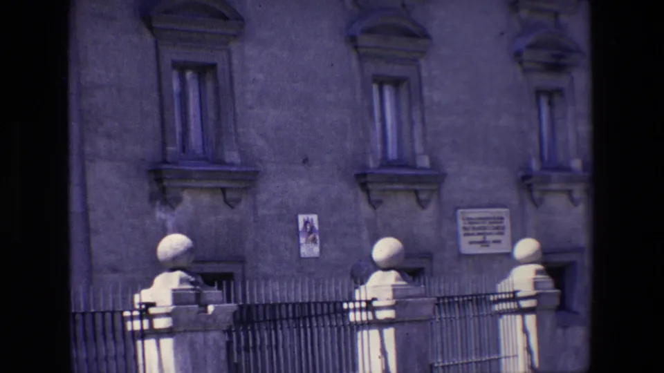 Wider 1967 Madrid home-movie view of an old facade with shuttered upper windows, a small poster, a wall plaque, iron fence rails, and stone posts along the street edge.