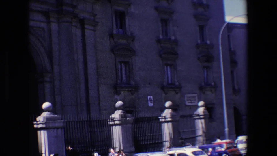 Wide 1967 Madrid street view showing the building exterior with an arched entry area, iron fence, rounded stone posts, wall signs, a small poster, and automobiles at the curb.
