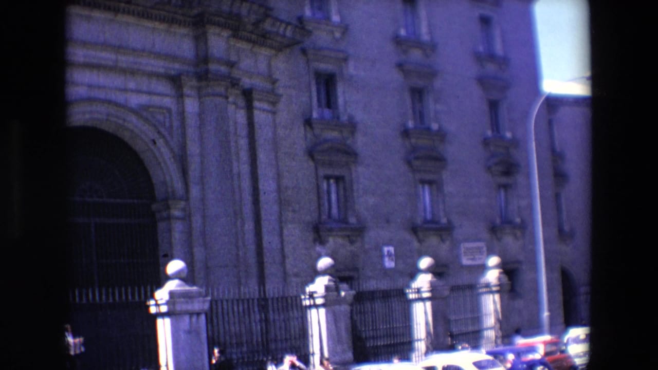 Stone building facade with arched gate, iron fence, pedestrians, and partial cars at street level.