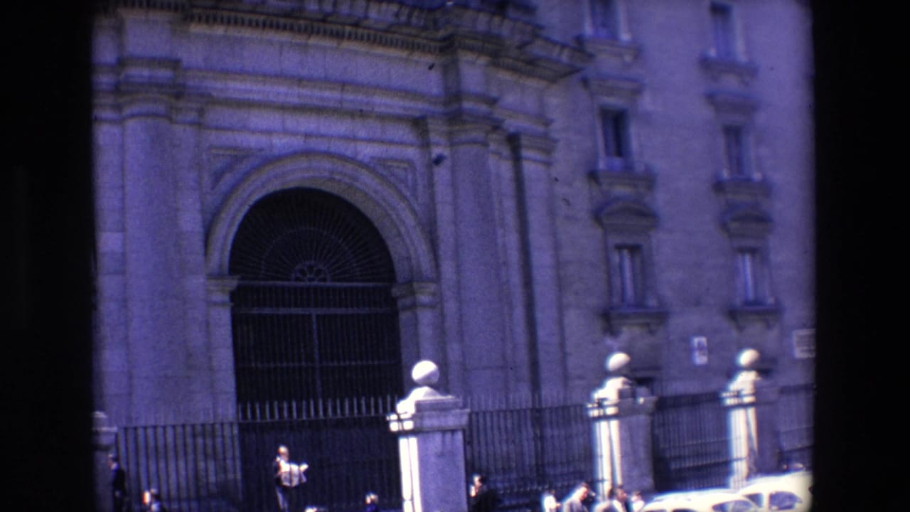 1967 Madrid frame with a monumental arched entrance, barred iron gate, stone facade details, pedestrians near the fence, and classic cars passing across the foreground.