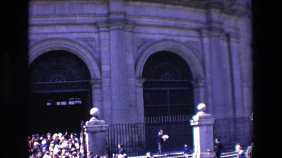 1967 Madrid home-movie view of multiple arched gated entrances and iron fencing, with people gathered near steps outside the large stone building.