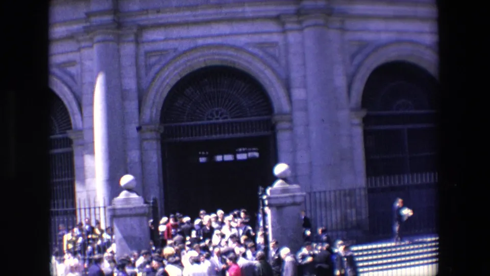 1967 Madrid frame showing a crowd near steps outside a large fenced stone building, with arched entrances and a narrow sunlit monument or post in the foreground.