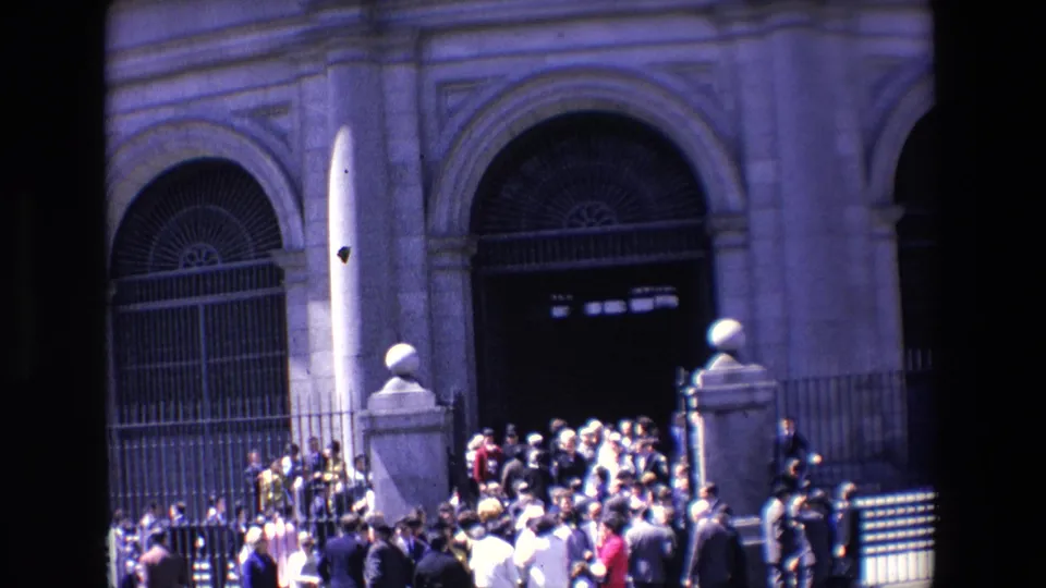 1967 Madrid home-movie frame showing people clustered near the steps and iron gates of a large arched stone building entrance.