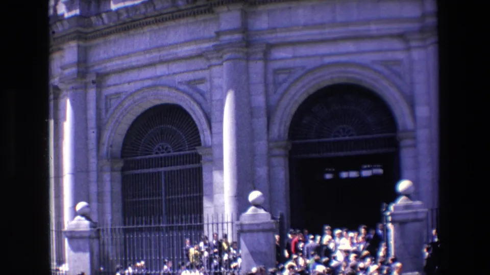 1967 Madrid home-movie frame showing a dense group near steps below a tall arched stone facade with barred gates.