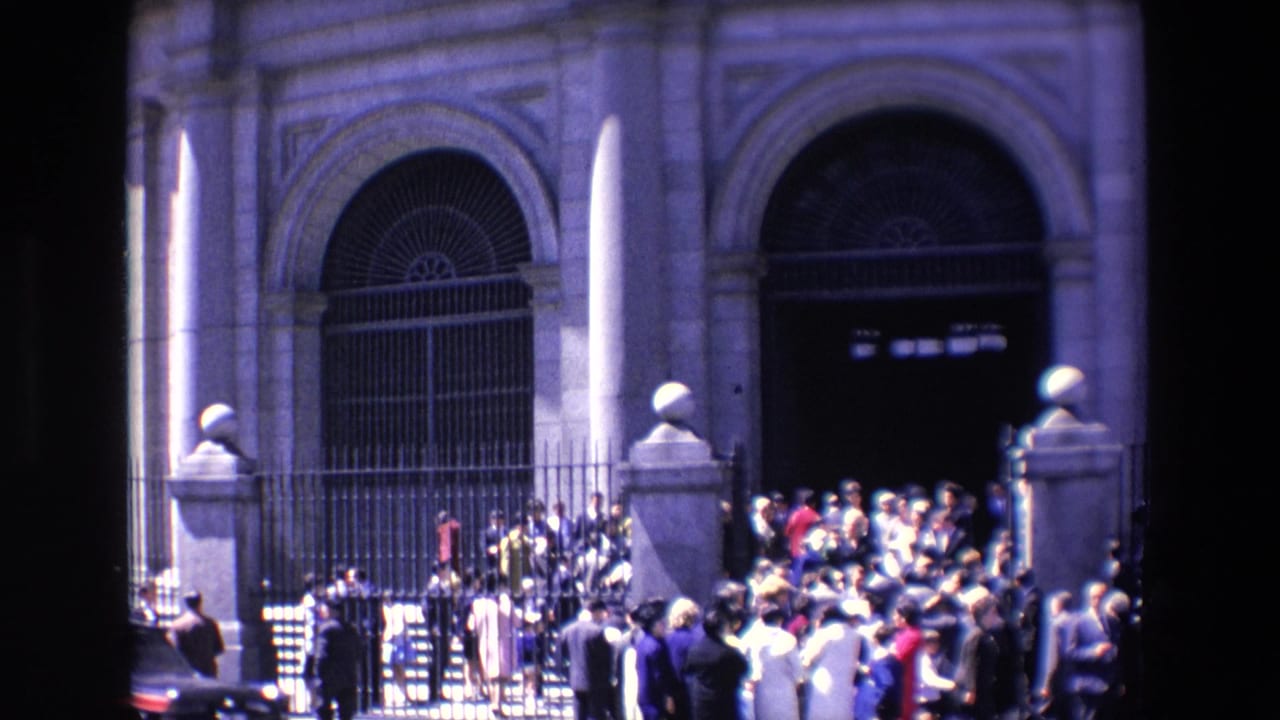 1967 Madrid home-movie frame showing pedestrians gathered near a wrought iron fence and arched stone building entrance, with strong sunlight and shadow.